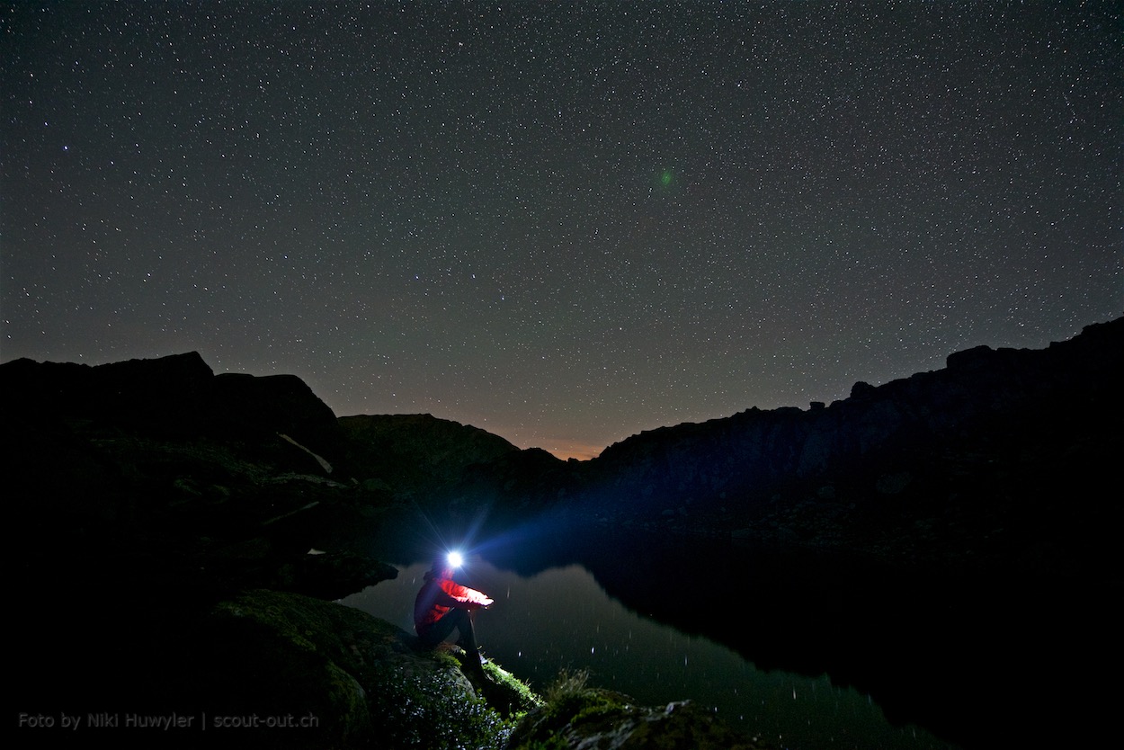 Nacht der Perseiden: Person mit Stirnlampe am Bergsee unter Sternen