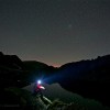 Nacht der Perseiden: Person mit Stirnlampe am Bergsee unter Sternen